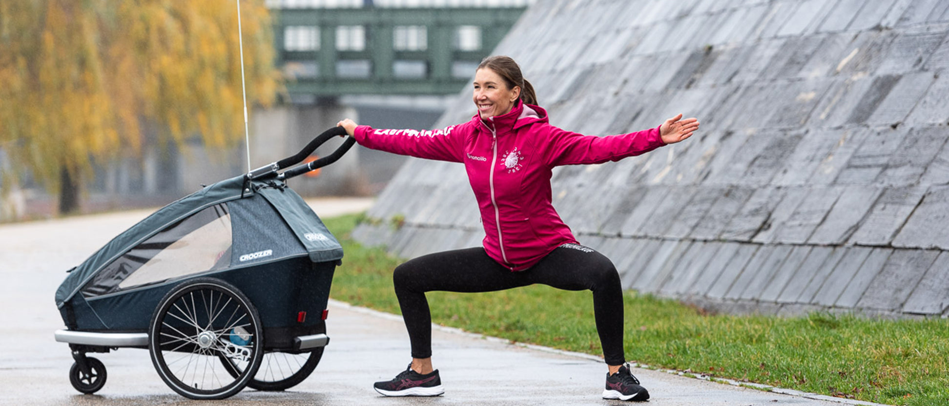 Woman performing wide squat with arms extended while holding Croozer bike trailer in autumn park setting
